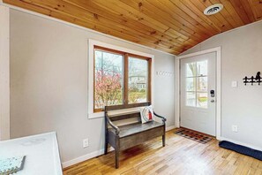 Ein Zimmer mit Holzboden, einer Bank, einer Tür mit Glasfenster und einem Fenster mit Blick auf ein Haus und Bäume.