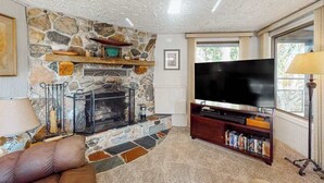 A living room with a stone fireplace, a flat-screen TV, and a bookshelf. - Riverfront Cabin with Stunning Views, Fire pit, Gas Grill & WIFI (Thompsonville)