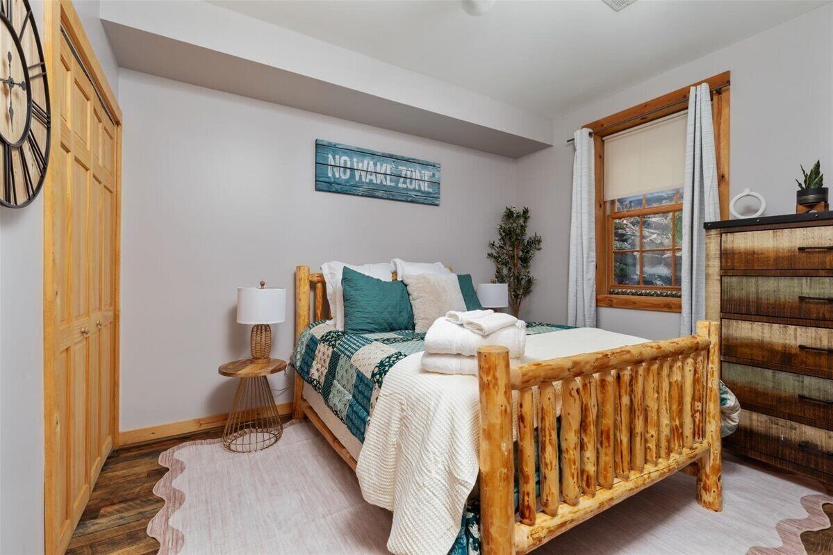 A bedroom with a wooden bed, a bedside lamp, a 'NO WAKE ZONE' sign, a window with curtains, and a wooden dresser.