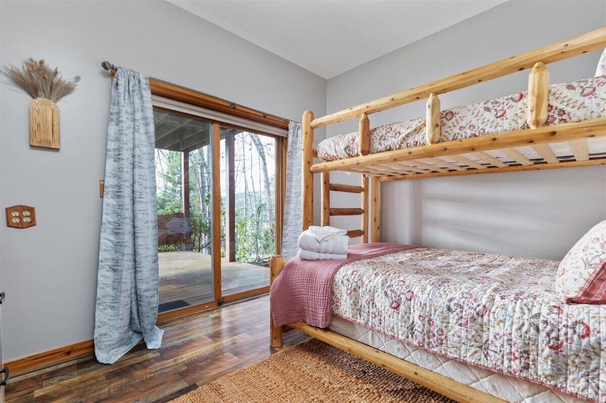 A bunk bed room with a wooden ladder, a checkered blanket, and a view through a glass door to an outdoor area.