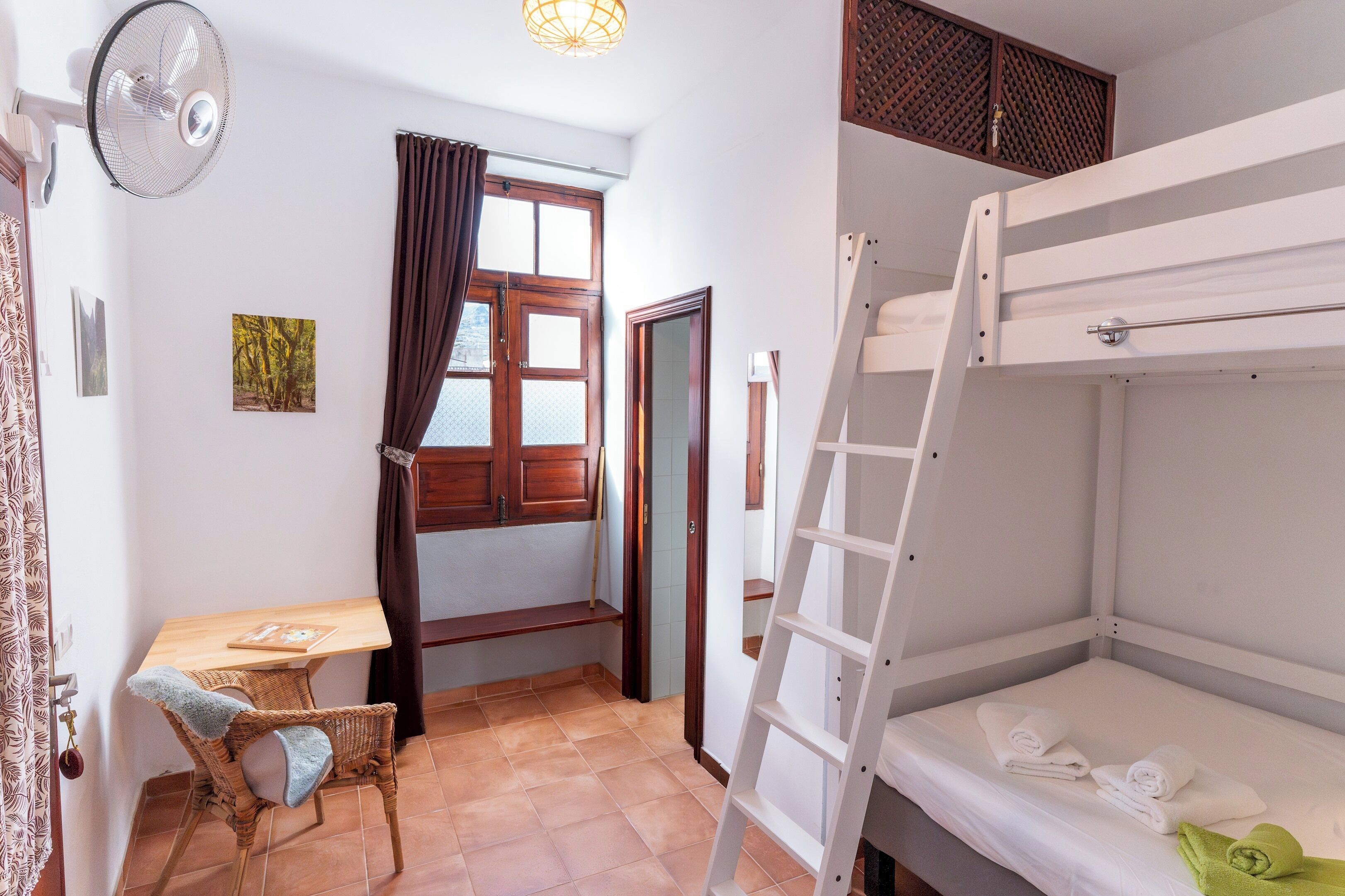 Una habitación con una cama nido, un escritorio con silla, un ventilador de pie y una ventana con cortinas.