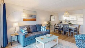 A living room with a sofa, coffee table, and dining area with chairs and a table. - Oceanfront Condo w/ Balcony & Amazing Beach Views (Ocean City)