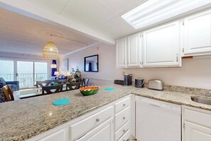 A modern kitchen with white cabinets, granite countertops, and a dining area with a table and chairs. - Oceanfront Condo w/ Balcony & Amazing Beach Views (Ocean City)
