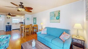 Ein Wohnzimmer mit blauem Sofa, einem Holzesstisch, Deckenventilatoren und einer Küche im Hintergrund.