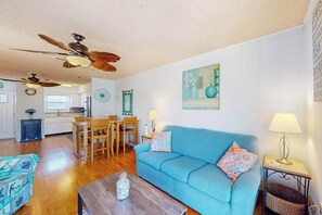 A living room with a blue sofa, wooden dining table, ceiling fans, and a kitchen in the background. - Water view condo with peaceful balcony, open layout, smart TVs, games, & W/D (Ocean City)