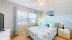 Ein Schlafzimmer mit Bett, Nachttisch, Lampe, Deckenventilator und Fenster mit Jalousien.