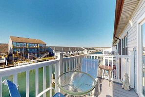 A balcony with a glass table and chairs, a white railing, and a view of a building across the water. - Water view condo with peaceful balcony, open layout, smart TVs, games, & W/D (Ocean City)