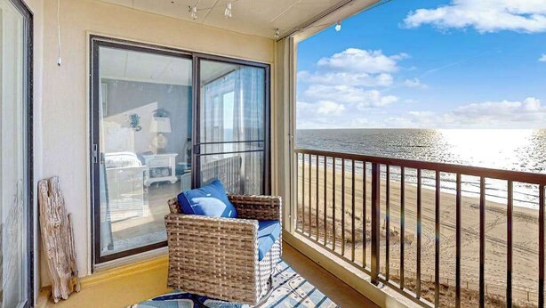 Een balkon met uitzicht op het strand, een rieten stoel met kussens, en een schuifdeur van glas.