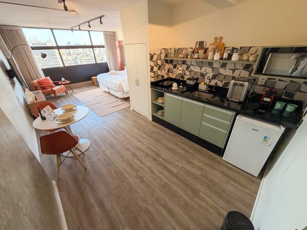 A compact hotel room with a kitchenette, a dining area with a table and chairs, a bed, and a view of the city through large windows. - Vem pro Mirante (São Paulo)