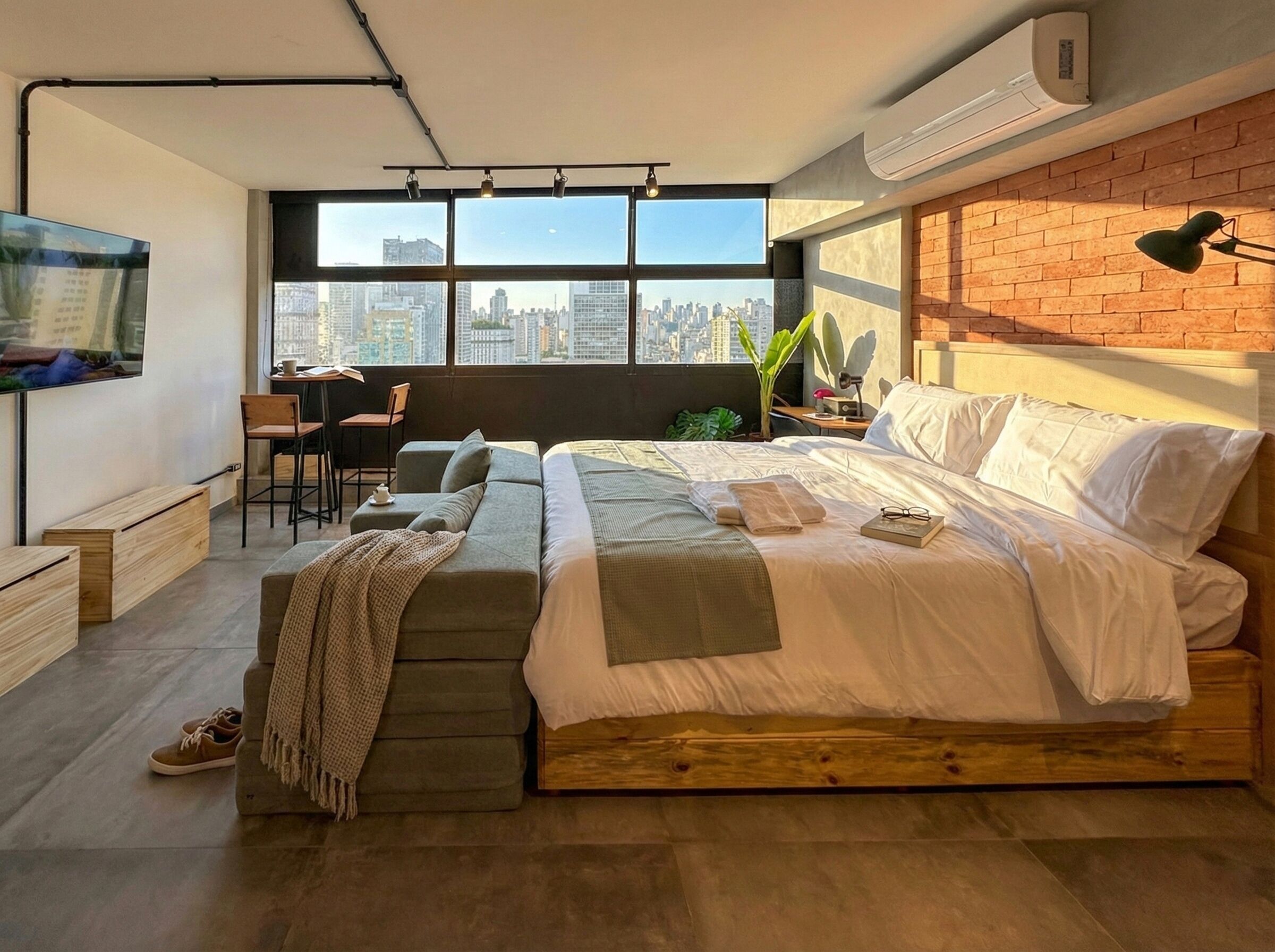 A modern bedroom with a city view, a sofa, a desk, and a bed with a brick wall.