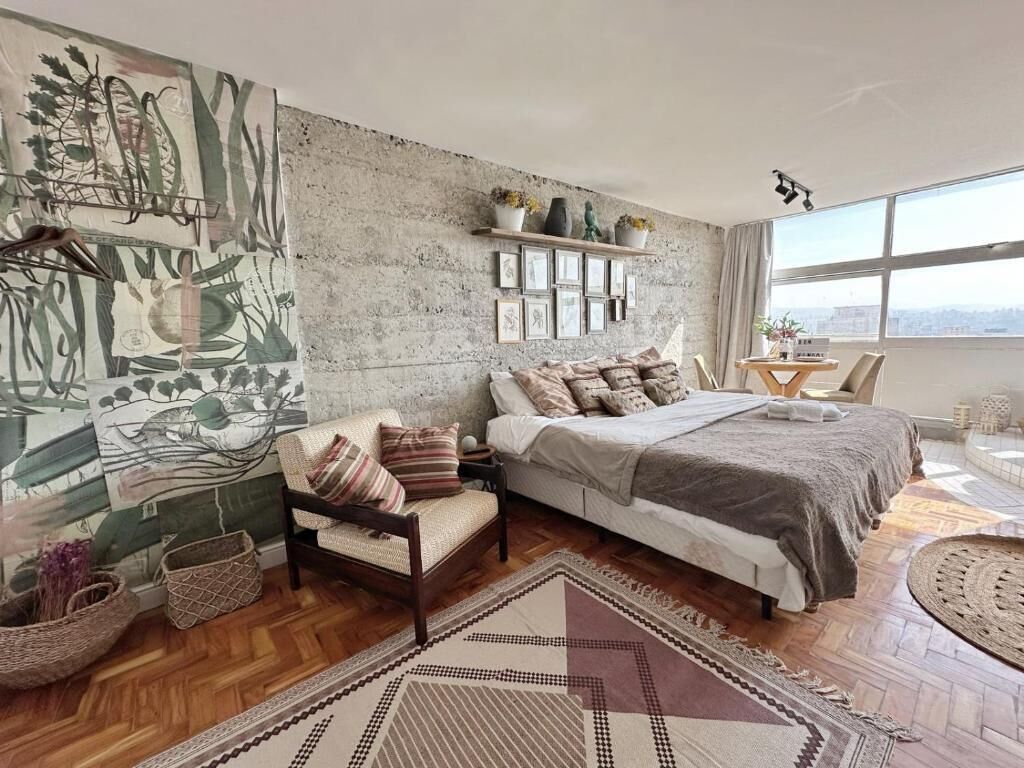 A bedroom with a large bed, a chair, a rug, a wall with artwork, and a window with a city view.