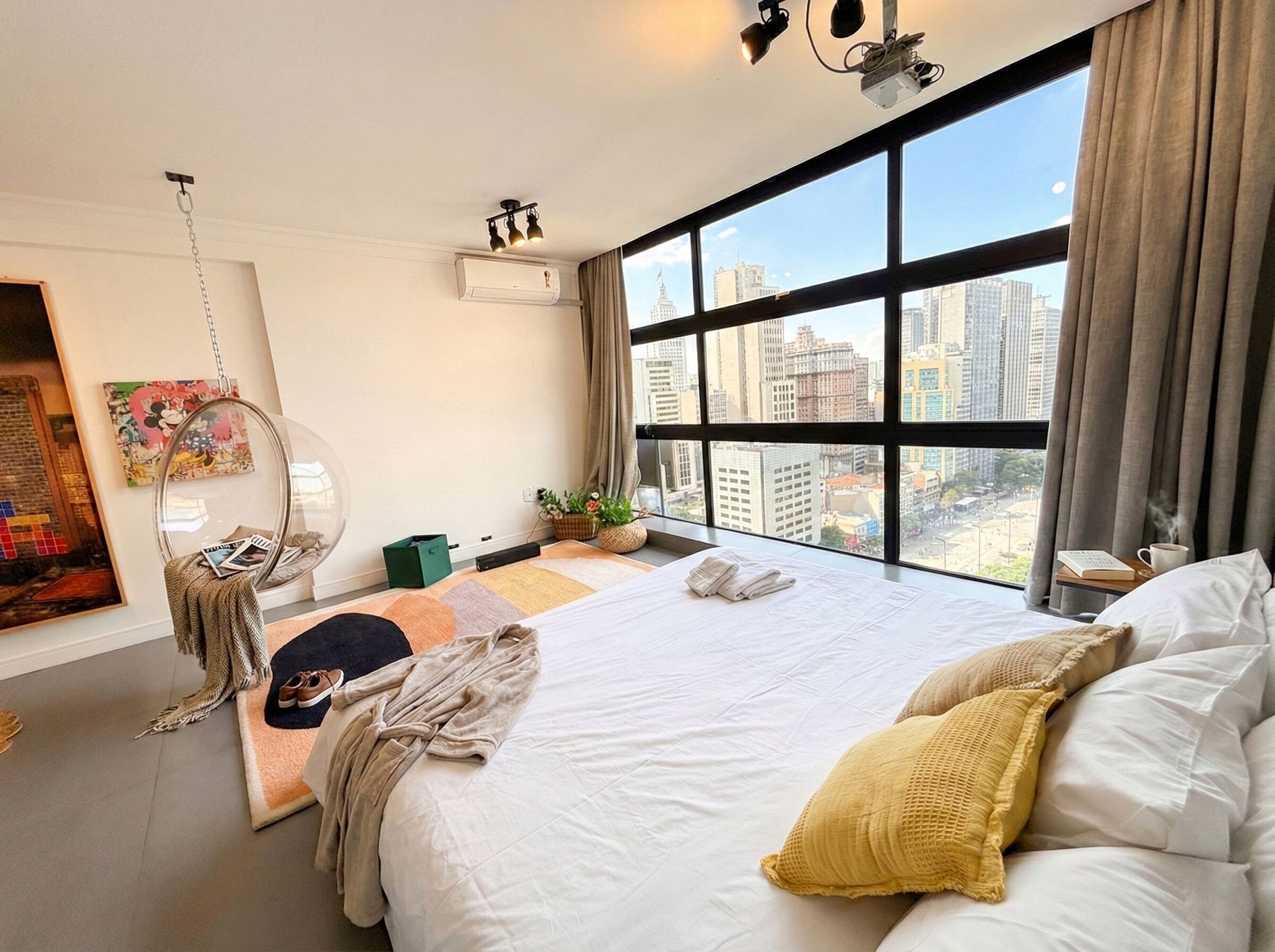 A modern hotel room with a large bed, a hanging chair, a view of the cityscape, and a window with curtains.