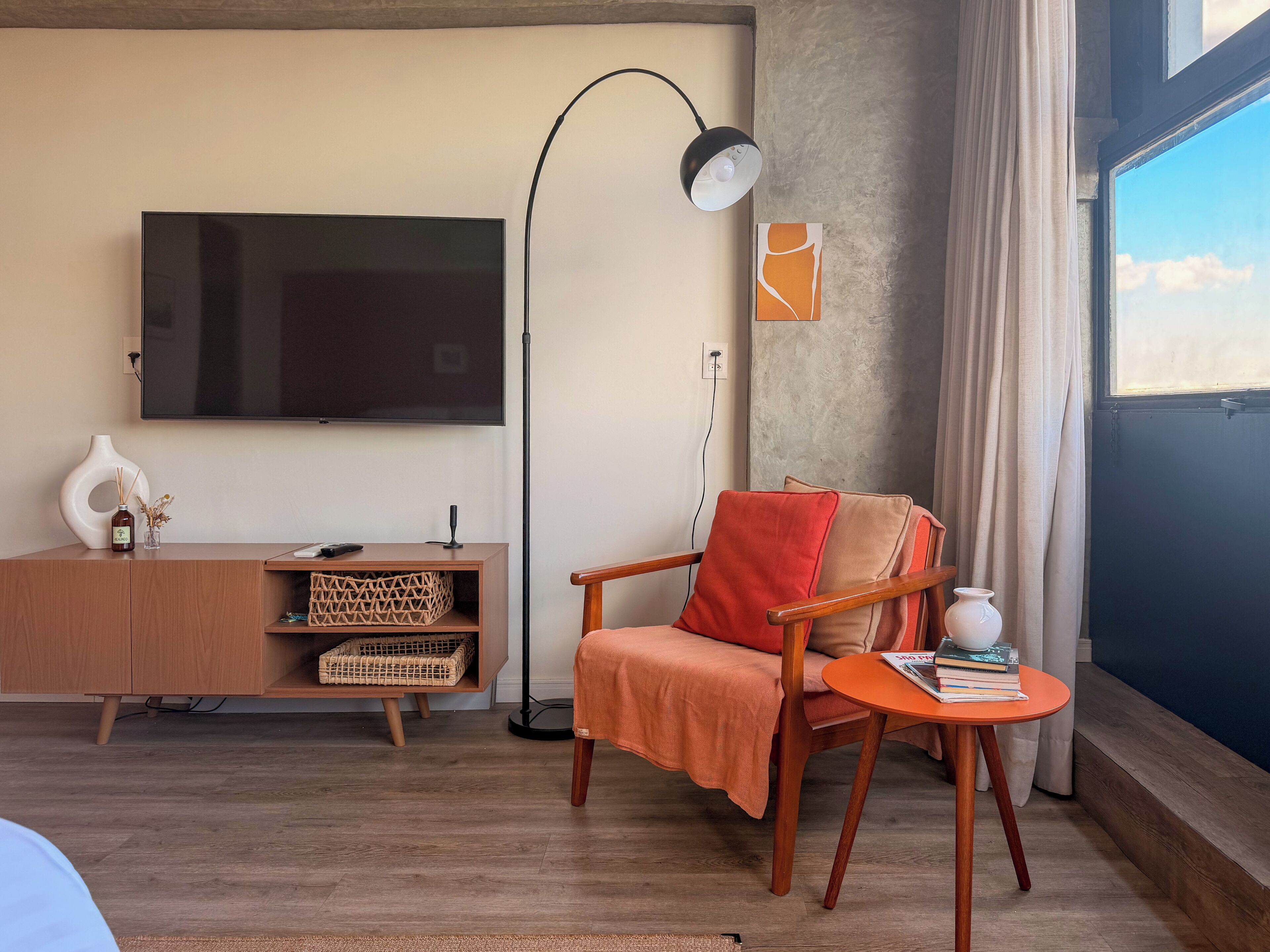 Una sala de estar con televisor de pantalla plana, un mueble de madera, una silla, una mesita con libros, una lámpara de pie y un ventanal con cortinas.