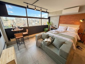 Una habitación moderna con un ventanal grande, una cama, un sofá, una mesita con bebidas y una silla.
