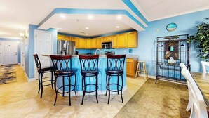 A kitchen with yellow cabinets, a blue island, and four black bar stools. - Bahia Vista II 304 (Ocean City)