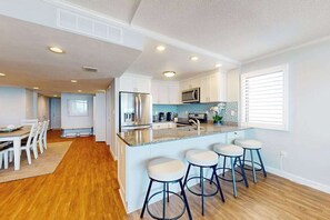 A modern kitchen with white cabinets, a stainless steel refrigerator, a microwave, and a bar area with stools. - Fountainhead Towers 1310 (Ocean City)