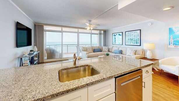 A modern kitchen with a granite countertop, stainless steel sink, and white cabinetry. - Fountainhead Towers 1310 (Ocean City)