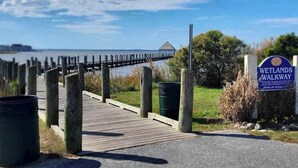Une promenade en bois menant à un plan d’eau, un panneau indiquant le sentier des zones humides et une structure visible au loin.
