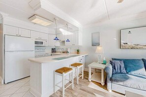 Eine moderne Küche mit weißen Schränken, einer zentralen Kochinsel mit Sitzgelegenheiten, einem kleinen Tisch mit Lampe und einem blauen Sofa im Wohnbereich.