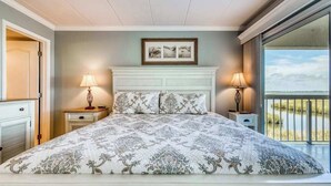 A bedroom with a large bed, a headboard, two bedside tables with lamps, a framed picture above the bed, and a view of the outdoors through a sliding door. - Triton's Trumpet B 101 (Ocean City)