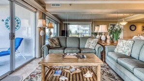 A living room with a sofa, coffee table, and lamps. There is a TV and a window with a view. - Triton's Trumpet B 101 (Ocean City)