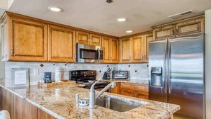A kitchen with wooden cabinets, a stainless steel refrigerator, a microwave, and a granite countertop. - Triton's Trumpet B 101 (Ocean City)