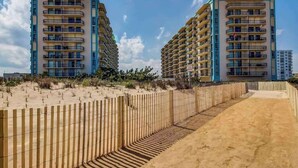 Un hotel frente a la playa con varios pisos, balcones y un paseo marítimo de arena.
