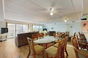 A spacious living area with a glass dining table surrounded by wicker chairs, a sofa, and a television on a white cabinet. - Triton's Trumpet C 303 (Ocean City)