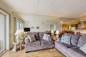 A living room with a sofa, two lamps, a television, and a dining area visible through a glass partition. - Triton's Trumpet C 303 (Ocean City)
