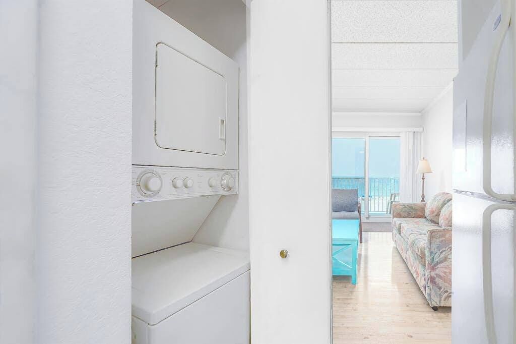 A laundry room with a washing machine and dryer, a door leading to a balcony with a view, and a living area with a sofa and a table.