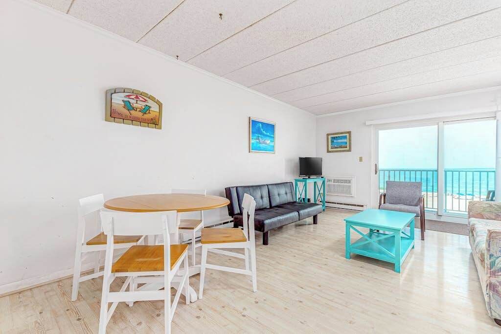 Une pièce avec un plancher en bois, des murs blancs, un canapé en cuir noir, une table basse turquoise et un téléviseur posé sur un meuble.