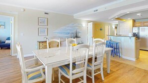 Un comedor con una mesa y sillas blancas, una cocina con refrigerador, y una sala de estar con sofá y un cuadro.