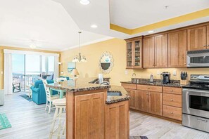 A kitchen with wooden cabinets, a granite countertop, stainless steel appliances, and a dining area with blue chairs. - Belmont Towers 610 (Ocean City)