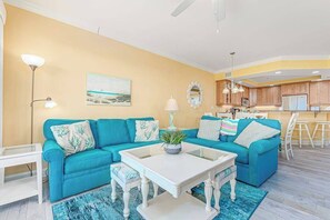 A living room with a teal sofa, a white coffee table, and a kitchen in the background. - Belmont Towers 610 (Ocean City)