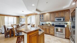 A modern kitchen with wooden cabinets, a central island, stainless steel appliances, and a dining area with a view. - Belmont Towers 801 (Ocean City)