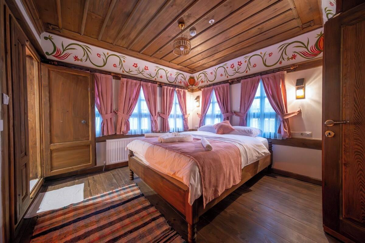 Quarto com painéis de madeira, uma cama, cortinas cor-de-rosa e um teto com estampas florais.