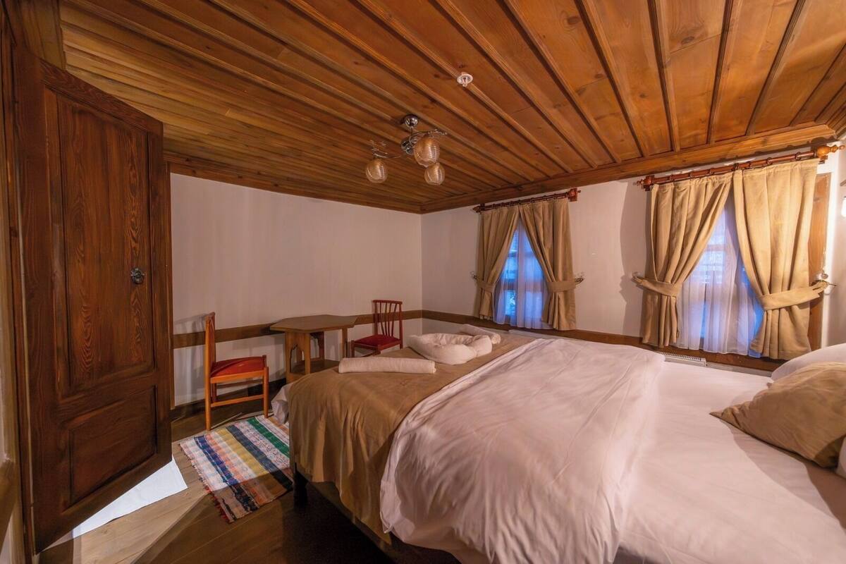 Quarto com painéis de madeira, uma cama, duas cadeiras, uma mesa pequena e uma janela com cortinas.