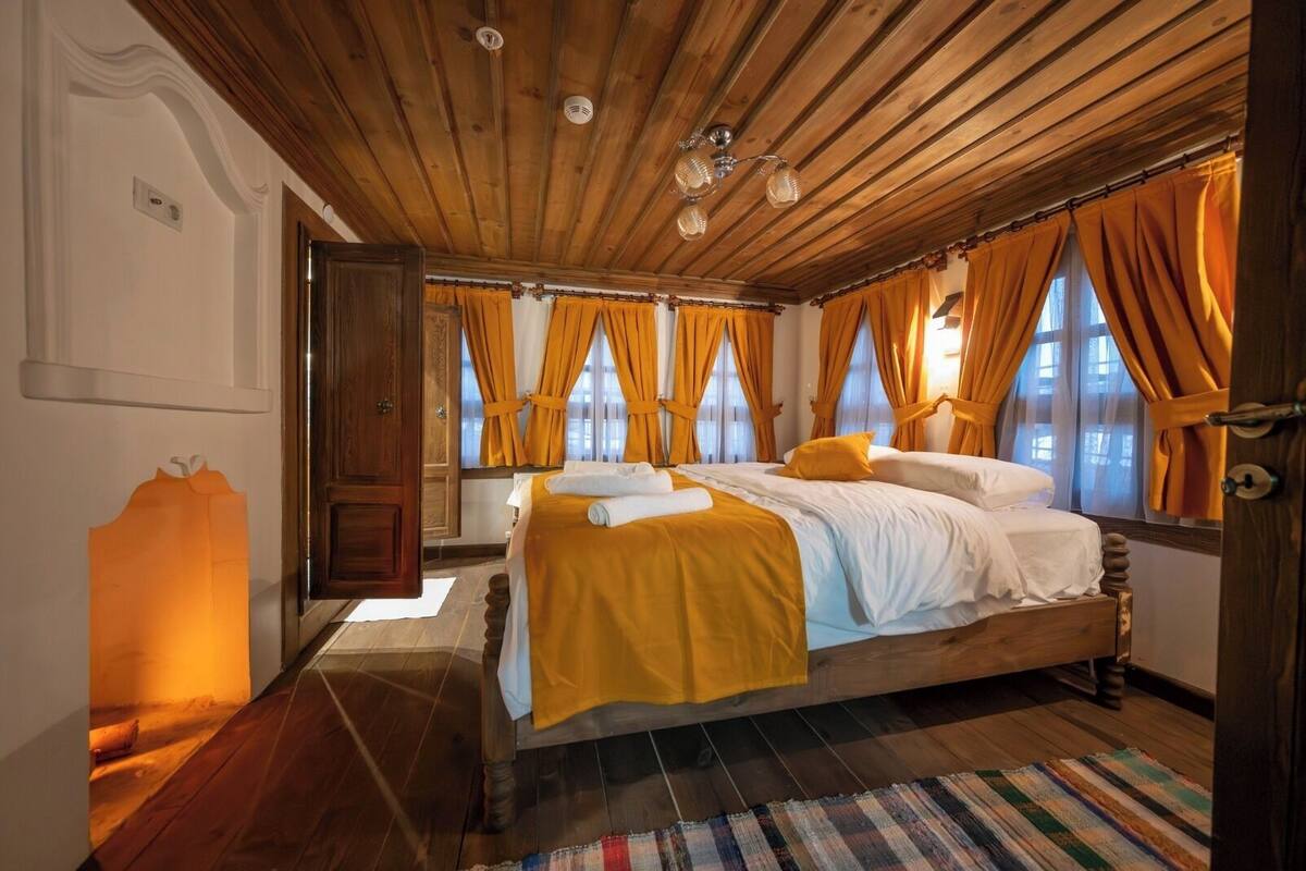 Quarto com teto de madeira, duas camas com roupa de cama branca e amarela, cortinas laranja e um tapete listrado.