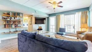 Ein Wohnzimmer mit einer Couch, Sesseln, einem Bücherregal, einem Flachbildfernseher, einem Deckenventilator und großen Fenstern.
