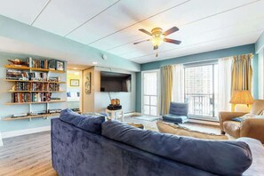 Ein Wohnzimmer mit einer Couch, Sesseln, einem Bücherregal, einem Flachbildfernseher, einem Deckenventilator und großen Fenstern.