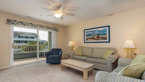 A living room with a ceiling fan, a sofa, a striped armchair, a coffee table, and a painting of a beach scene. - Baywatch I - 201B (Ocean City)