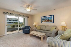 A living room with a ceiling fan, a sofa, a striped armchair, a coffee table, and a painting of a beach scene. - Baywatch I - 201B (Ocean City)