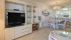 A modern living room with a flat-screen TV, glass shelves, a dining area with white chairs, and a kitchen with white cabinets. - Anchorage  304 (Ocean City)