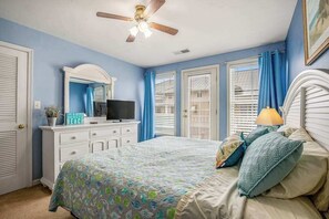 Ein Schlafzimmer mit einem Bett, einem Fernseher auf einer Kommode, einem Deckenventilator und einem Fenster mit Vorhängen.