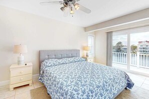 A bedroom with a bed, a nightstand, a lamp, a ceiling fan, and a large window with a view. - Sunset Harbour townhouse with balcony, & central AC (Ocean City)