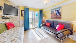 Una stanza con un letto, un divano, una TV, una ventola a soffitto e un balcone con tende azzurre.