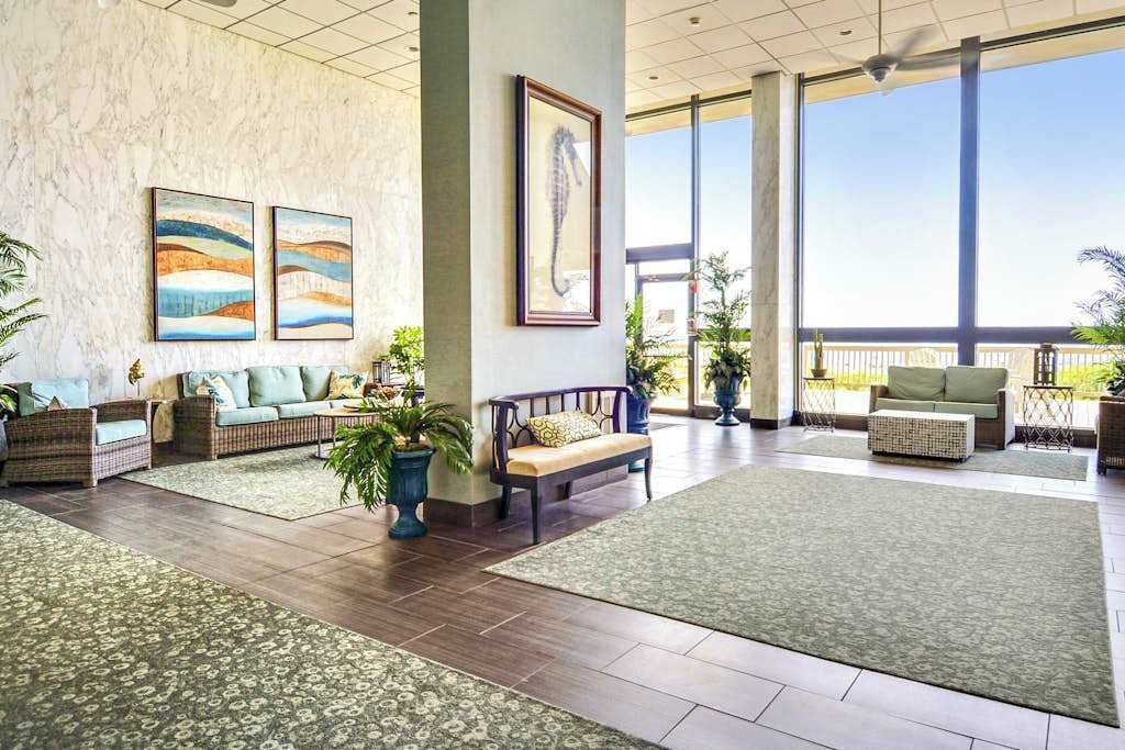 Eine geräumige Lobby mit hoher Decke, großen Fenstern und Blick ins Freie. Es gibt mehrere Sitzbereiche mit Kissen und Topfpflanzen.