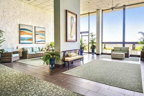 Eine geräumige Lobby mit hoher Decke, großen Fenstern und Blick ins Freie. Es gibt mehrere Sitzbereiche mit Kissen und Topfpflanzen.