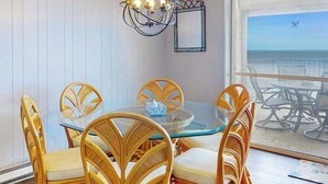 A dining area with a glass table and wicker chairs, a chandelier, and a view of the beach through a large window. - Calypso 705 (Ocean City)