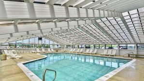 Une piscine intérieure dotée d’un haut plafond, de panneaux de verre transparent et d’espaces baignés de lumière naturelle, aménagés avec des chaises longues.